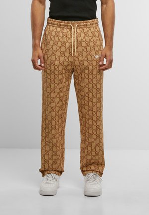 Brown patterned sweatpants with an elastic waistband and drawstring, featuring a repeated logo design, paired with white sneakers.