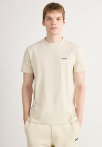 Beige színű, rövid ujjú póló sima anyagból, kerek nyakkivágással és egy kis fekete "BOSS" logóval a bal mellrészen.