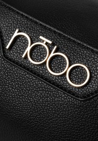 Superficie in pelle nera testurizzata con logo "nōbo" in tono oro e caratteri distintivi. Nota gli accenti cuciti lungo i bordi.