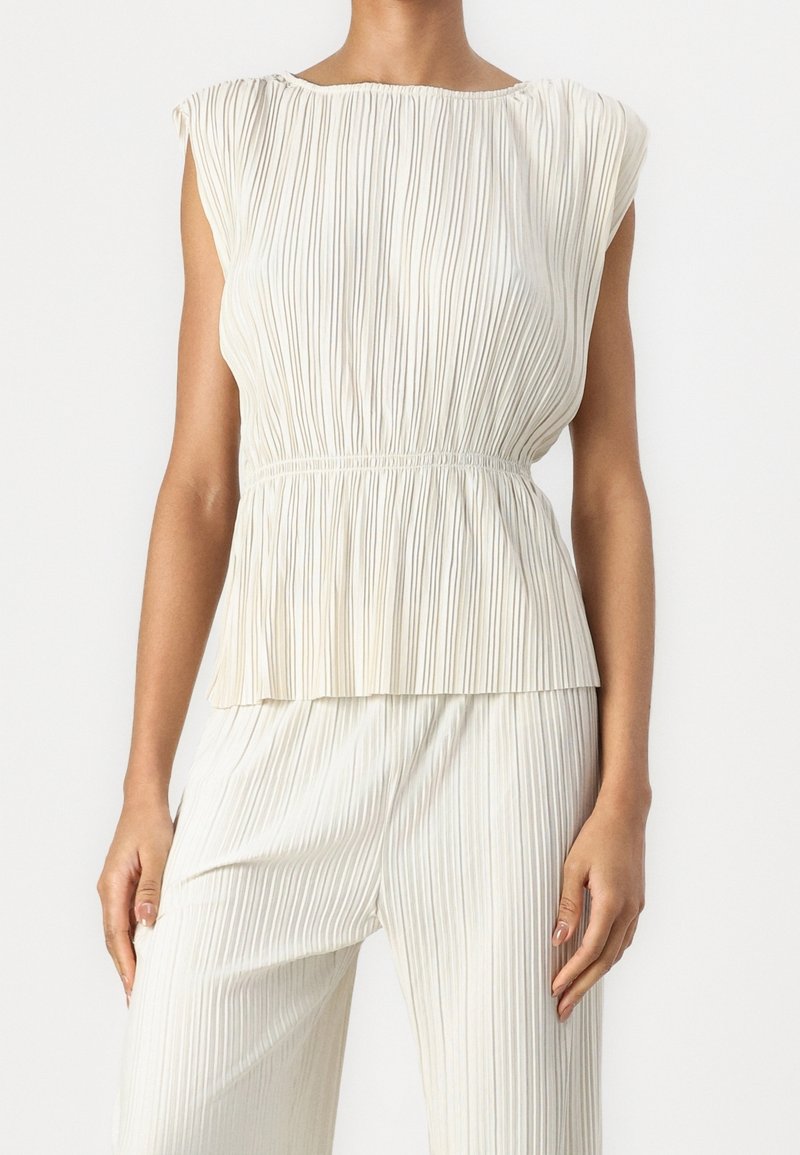 Femme portant un haut sans manches blanc à plis verticaux et un pantalon palazzo plissé assorti sur un fond uni.