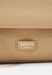 Borsa in pelle beige con finitura testurizzata, con un'etichetta Lancel Paris in oro e beige cucita sul davanti.
