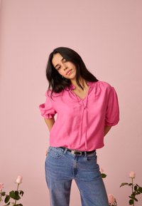 Jeune femme aux longs cheveux noirs portant un chemisier rose vif et un jean bleu, debout devant un fond rose avec des roses de chaque côté.