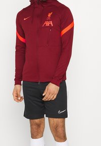 Bordeauxfarbene Zip-Jacke mit Kapuze. Mit dem Liverpool FC-Logo und "AXA" in Orange. Verfügt über eine Reißverschlusstasche und kontrastierende orangefarbene Streifen an den Ärmeln.