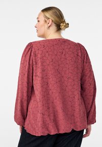 Texturerad, vinröd långärmad blus med ett blommönster, med samlade axlar och en vågad nederkant. Bakvy visas.