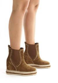 Brązowe zamszowe botki za kostkę z elastycznymi panelami bocznymi, ćwiekowanymi akcentami oraz teksturowaną podeszwą na koturnie, charakteryzujące się miękkim wykończeniem i kontrastowymi elementami wzorniczymi.