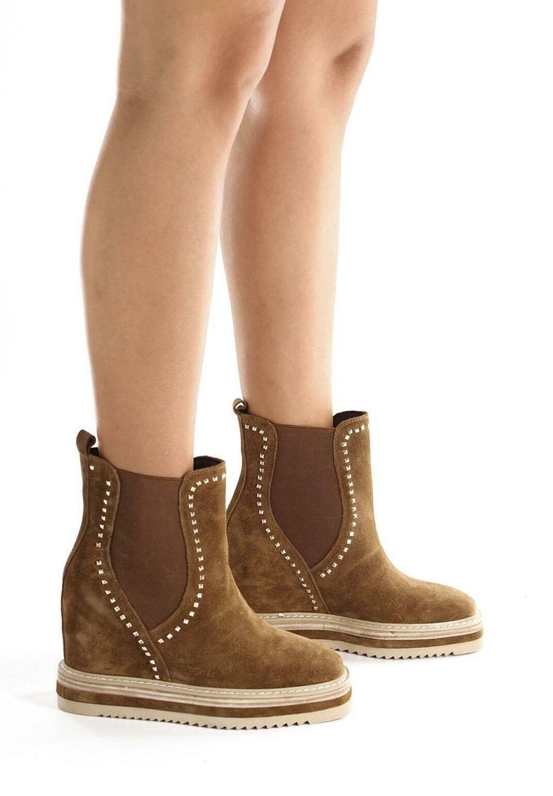 Brązowe zamszowe botki za kostkę z elastycznymi panelami bocznymi, ćwiekowanymi akcentami oraz teksturowaną podeszwą na koturnie, charakteryzujące się miękkim wykończeniem i kontrastowymi elementami wzorniczymi.