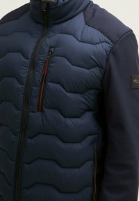 Doudoune marine avec texture matelassée hexagonale, col haut et manches foncées contrastantes ; dotée d'une fermeture éclair rouge et d'un patch logo.