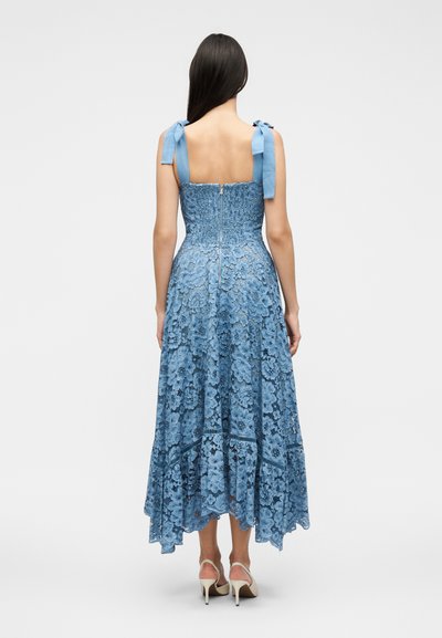 Γυναίκα με μπλε midi φόρεμα από δαντέλα, με ραμμένη κοντή μπλούζα, δέστρες στους ώμους, φερμουάρ πίσω και ασύμμετρο τελείωμα, φοράει λευκά σανδάλια με ψηλό τακούνι.