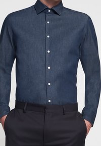 Mann in einem dunkelblauen Hemd mit Knöpfen, das in schwarze Hose gesteckt ist, mit Händen in den Taschen, von Hals bis Hüften gezeigt.