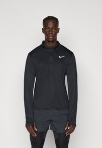 Svart långärmad atletisk topp med halvkedja, med en vit Nike-logotyp på vänstra bröstet, tillverkad av lätt material.