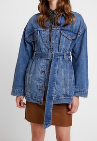 Blå denimjacka med bälte i midjan, med stora fickor, knappstängningar och tydlig sömnad. Bärs över en mönstrad skjorta.