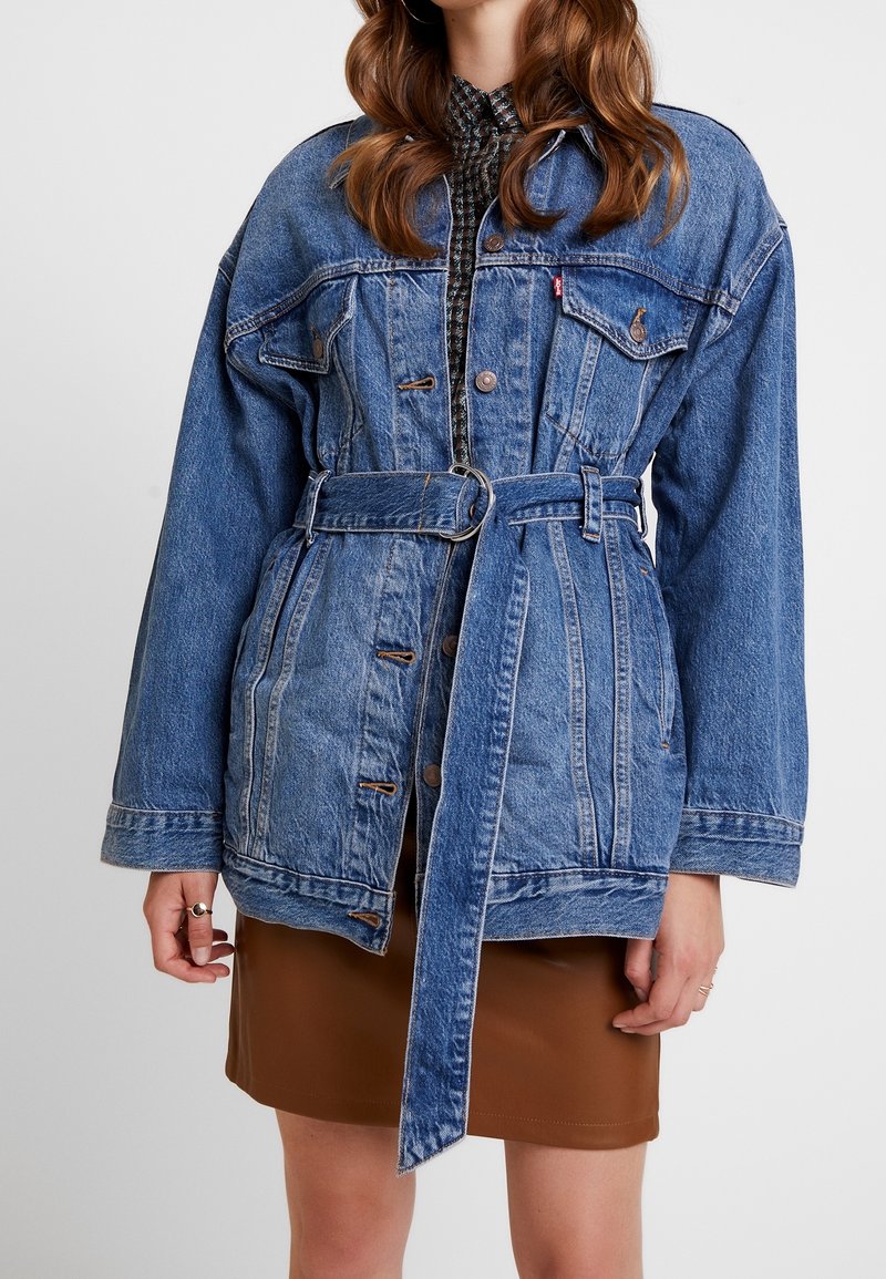 Blå denimjacka med bälte i midjan, med stora fickor, knappstängningar och tydlig sömnad. Bärs över en mönstrad skjorta.