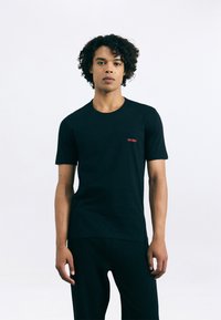 Tricou din bumbac negru, cu guler rotund și mâneci scurte, având un mic logo roșu "HUGO" pe partea stângă a pieptului. Textură netedă.