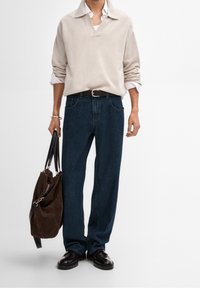 Pull-over en tricot beige avec un col en V, jeans bleu marine à jambes larges, ceinture noire, sac en daim marron et chaussures noires. Design simple, motifs minimalistes.