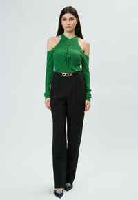 Grünes geripptes Off-Shoulder-Oberteil mit Schnürdetails, kombiniert mit hochgeschnittenen schwarzen Hosen und einem Kettenakzent-Gürtel. Schwarze spitz zulaufende Schuhe.