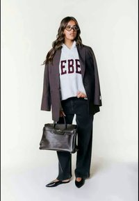 Grå hettegenser med teksten "EBE", svart blazer, mørke vide bukser, svarte flate sko, og en brun håndveske. Har langt, bølget hår og briller.