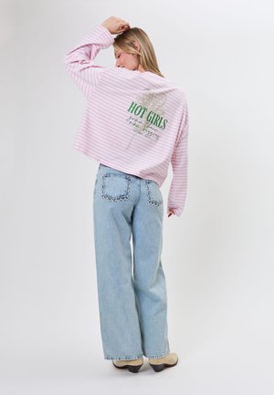 Kvinde i lyserød, stribet langærmet skjorte med grøn tekst på ryggen, lyseblå jeans med bølget lomme-detaljer og beige sko.