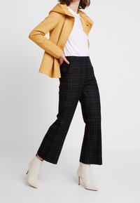 Gul tekstureret jakke med hætte, kombineret med sorte ternede bukser med brune linjer og hvide ankelstøvler med hæl.