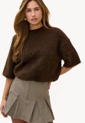 Femme aux cheveux longs portant des boucles d'oreilles créoles dorées, un pull à manches courtes marron duveteux, et une mini-jupe plissée à motif pied-de-poule sur fond blanc.