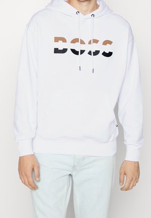 Person iført en hvid hættetrøje med "BOSS" tekst i brun, hvid og sort tværs over brystet, kombineret med lyseblå jeans.