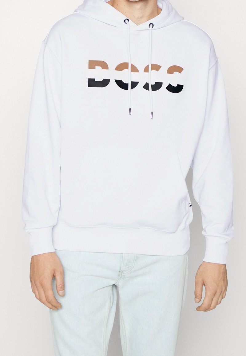 Personne portant un sweat à capuche blanc avec le texte "BOSS" en marron, blanc et noir sur la poitrine, associé à un jean bleu clair.