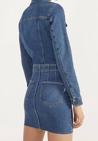 Kvinna klädd i en åtsittande blå denimjacka och matchande högmidjad denimkjol med bakfickor, ståendes mot en enfärgad bakgrund.