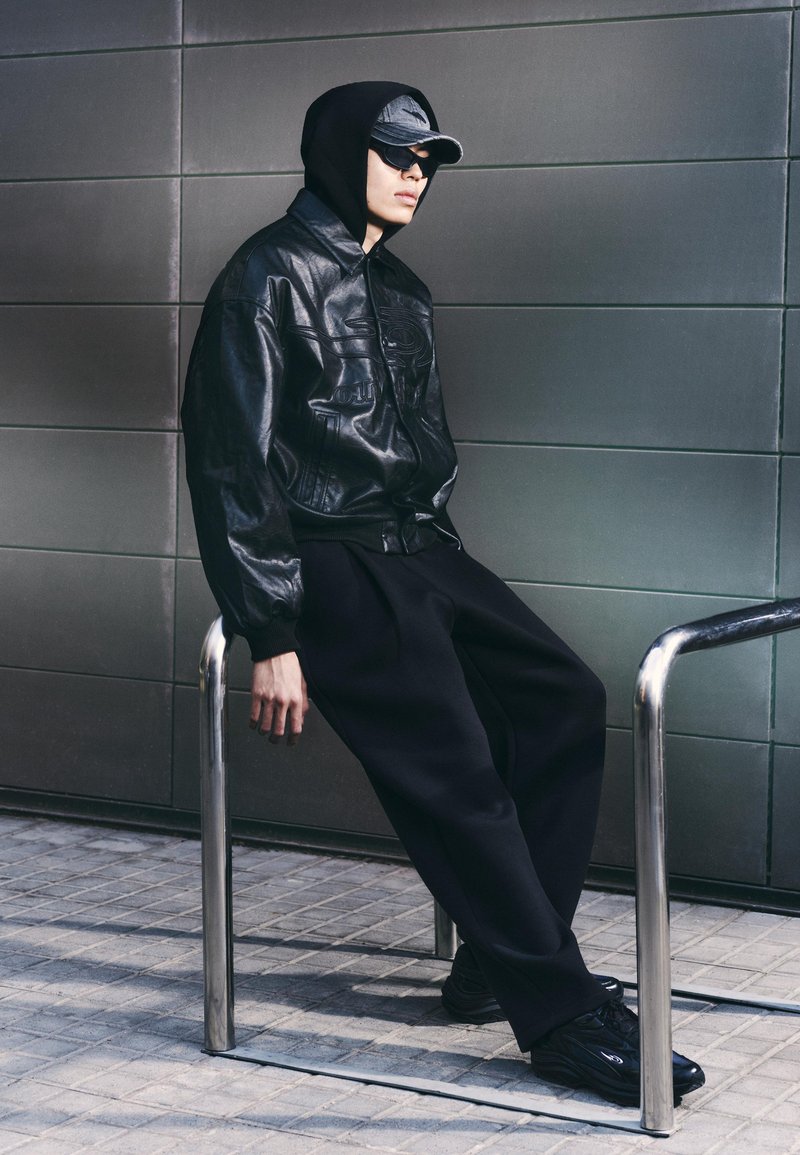 Fiatal fekete bőrkabátot, fekete kapucnis pulóvert, sapkát és napszemüveget viselő személy, aki egy fém kerékpártartón támaszkodik vissza egy csempézett fal előtt.