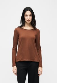 Γυναίκα με μακριά σκούρα μαλλιά που φορά καφέ πουκάμισο με μακριά μανίκια και μαύρο παντελόνι, στέκεται μπροστά σε απλό ανοιχτό γκρι φόντο.