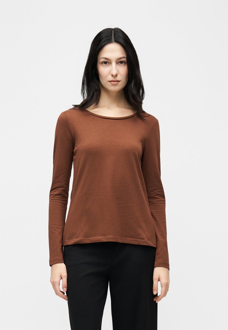 Γυναίκα με μακριά σκούρα μαλλιά που φορά καφέ πουκάμισο με μακριά μανίκια και μαύρο παντελόνι, στέκεται μπροστά σε απλό ανοιχτό γκρι φόντο.