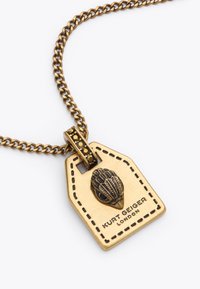 Collana in bronzo con un pendente a forma di etichetta, caratterizzato da un design a conchiglia, testo inciso "Kurt Geiger London" e una catena texturizzata.