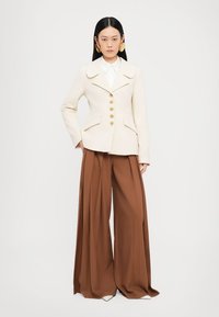 Blazer beige ajustado con cuello chal, botones dorados y dos bolsillos frontales, combinado con pantalones anchos marrones y pendientes redondos de gran tamaño.