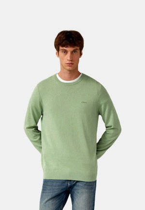 Młody mężczyzna z krótkimi brązowymi włosami, ubrany w jasnzięly sweter z okrągłym dekoltem na białej koszuli i niebieskich dżinsach, stoi z rękami za plecami.