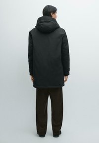 Sort hættejakke med afslappet pasform, mat finish og minimalistisk design. Lommer foran; båret med brune bukser.