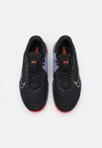 Een paar zwarte atletiekschoenen van mesh met paarse accenten, rode zolen, zwarte veters en witte Swoosh-logo's, gefotografeerd van bovenaf op een witte achtergrond.