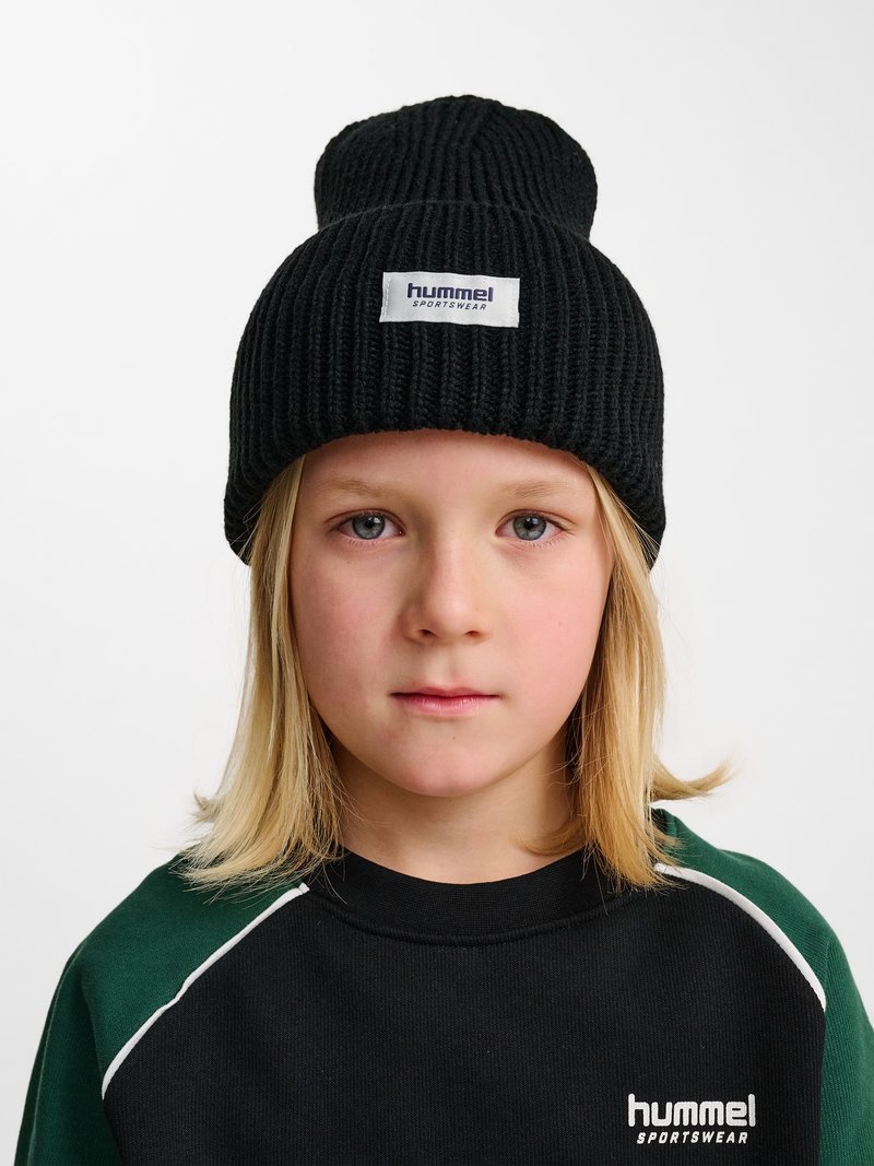Barn med lige blondt hår bærer en sort strikket hue og sort og grøn sportstop med "hummel sportswear" logo.