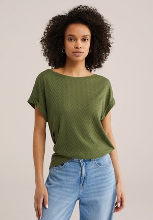 Göndör hajú nő zöld, texturált, rövid ujjú felsőt visel, amelyet világoskék farmerral tűrt be, és egyszerű háttér előtt áll.