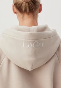 Plava žena sa kosom u punđi nosi bež duksericu s natpisom "LeGer" izvezenim na leđima kapuljače.
