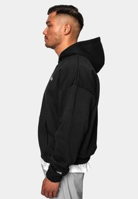 Hoodie noir en coton, présentant une poche avant, une capuche avec cordon de serrage et des poignets côtelés. Détail du logo blanc près de l'épaule.