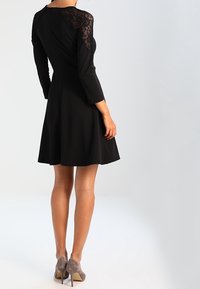 Czarna sukienka z rękawami 3/4 z koronki, dopasowaną górą i rozkloszowanym dołem. Noszona z szarymi szpilkami o gładkiej fakturze i klasycznym designie.
