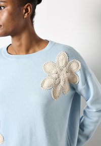 Jersey de punto azul claro con un gran acento de flor de ganchillo en color crema en el hombro izquierdo. Cuello redondo y corte relajado.
