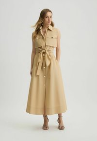 Abito senza maniche beige con vita con cintura, chiusura a bottoni, e due tasche sul petto, caratterizzato da una gonna fluida e un tessuto testurizzato.
