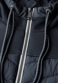 Marinblå jacka med kviltad textur, kontrasterande dragkedja, snörning i huvan och grå detaljer längs fram.