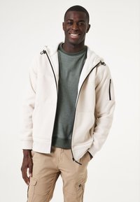 Un hoodie zippé gris clair avec une doublure en molleton doux, associé à un pull vert foncé et un pantalon cargo beige, avec une poche zippée sur le côté.