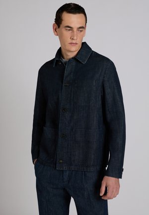 Mężczyzna w ciemnoniebieskiej kurtce dżinsowej z guzikami i dopasowanych dżinsowych spodniach, ręce w kieszeniach, patrzący w dół na bok.