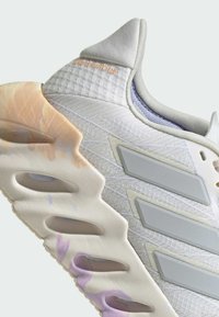 Sneaker bianca con upper testurizzato, tre strisce grigie e una suola in gomma colorata con ritagli e una punta liscia.