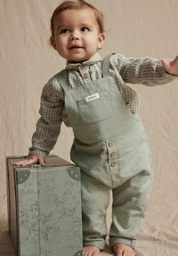 Salopette en coton vert clair avec des bretelles réglables et des fermetures à bouton, assortie à une chemise à carreaux. Une boîte grise décorée est à proximité.