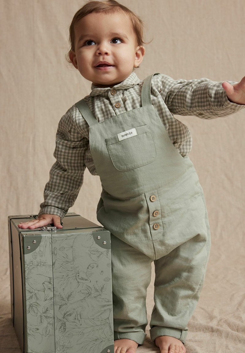 Salopette en coton vert clair avec des bretelles réglables et des fermetures à bouton, assortie à une chemise à carreaux. Une boîte grise décorée est à proximité.