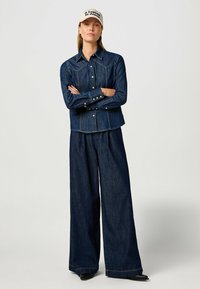 Giubbotto in denim scuro con chiusura a bottoni e colletto classico, abbinato a pantaloni in denim scuro a gamba larga, entrambi con dettagli di cucitura sottili.