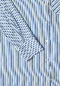 Chemise bleu clair avec des rayures verticales bleu marine et blanches, des boutons blancs et un poignet boutonné sur la manche gauche.