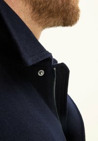 Musta villakangastakki korkealla kauluksella, jossa on vetoketjusuljenta ja napinappi. Sileä pinta ja rakenteellinen muotoilu korostavat kaulusta.